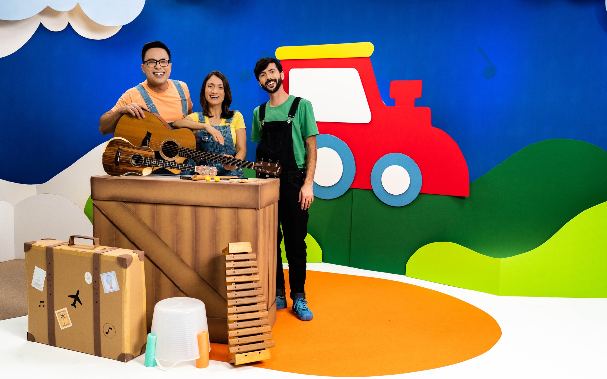 Daniel, Marla e André, do Viagem Musical, atrás de uma mesa com violão e instrumentos de percussão